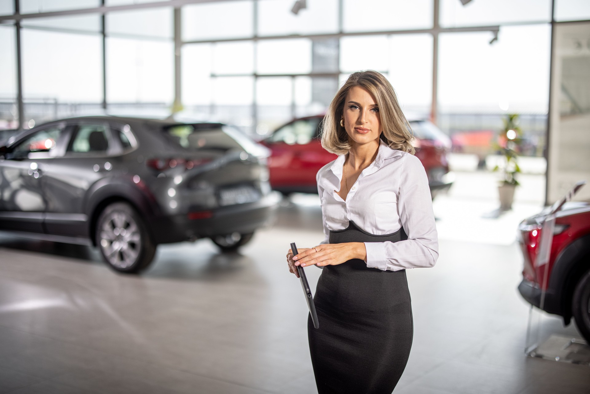 Young well dressed sales woman standing in car dealership saloon with tablet